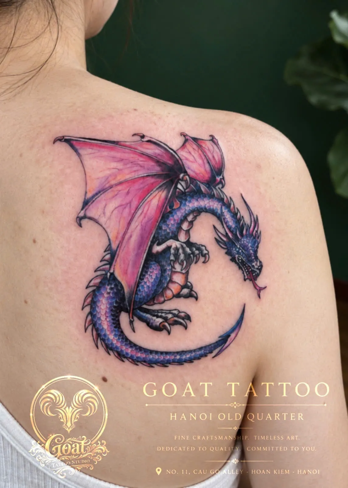 A full-color fantasy dragon with pink wings coiled across the shoulder blade, by GOAT Tattoo Studio Hanoi.