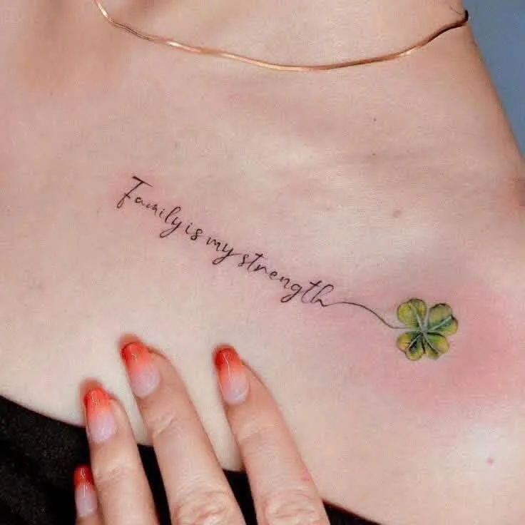 A cursive Family Is My Strength script leading into a green four-leaf clover, tattooed on the collarbone, by GOAT Tattoo Studio Hanoi.