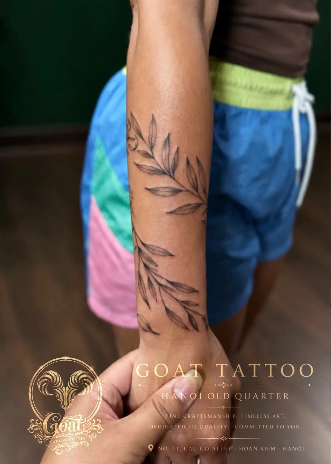 A fine line leaf branch wrapping around the forearm, by GOAT Tattoo Studio Hanoi.