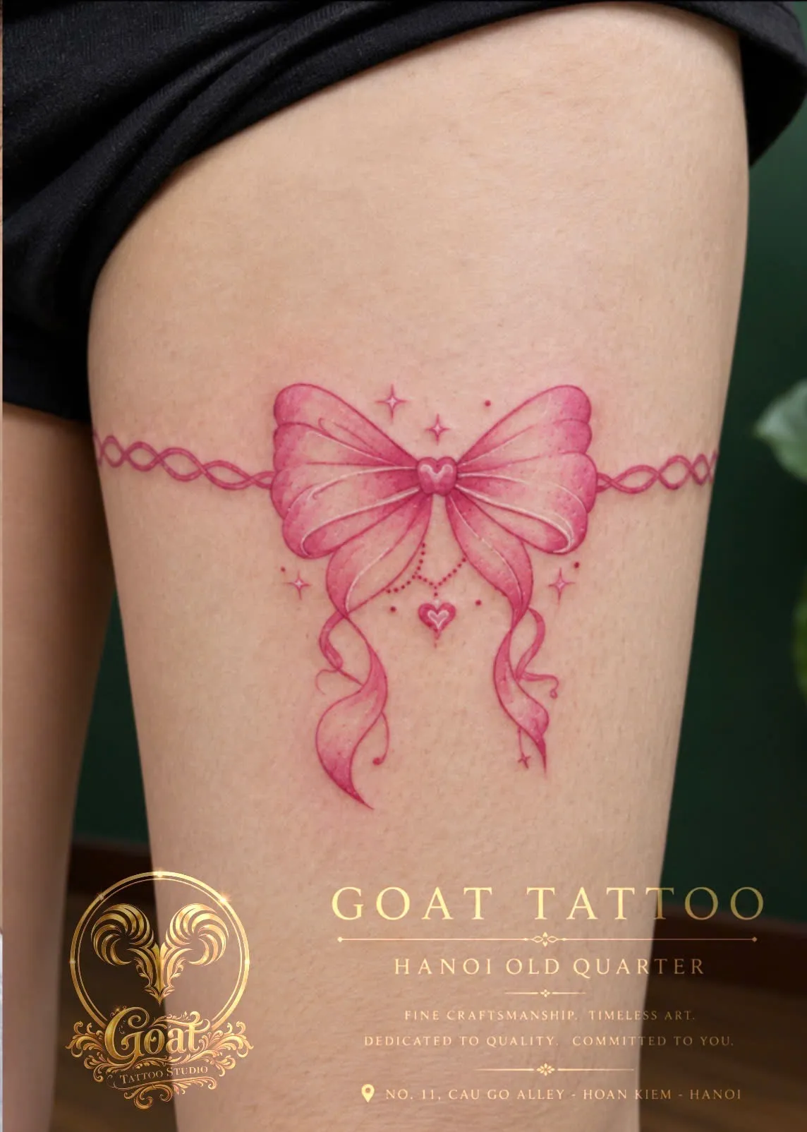 A pink ribbon bow tattoo styled like a garter band around the thigh, by GOAT Tattoo Studio Hanoi.