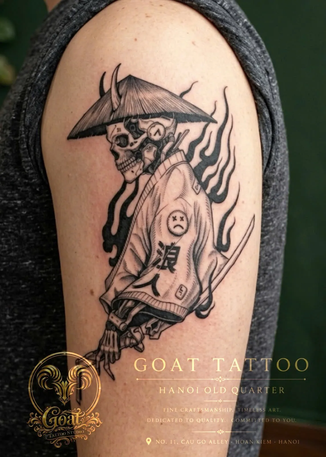 A black and grey illustrative tattoo of a skeletal ronin samurai in a conical hat with kanji lettering, on the upper arm, by GOAT Tattoo Studio in Hanoi's Old Quarter.