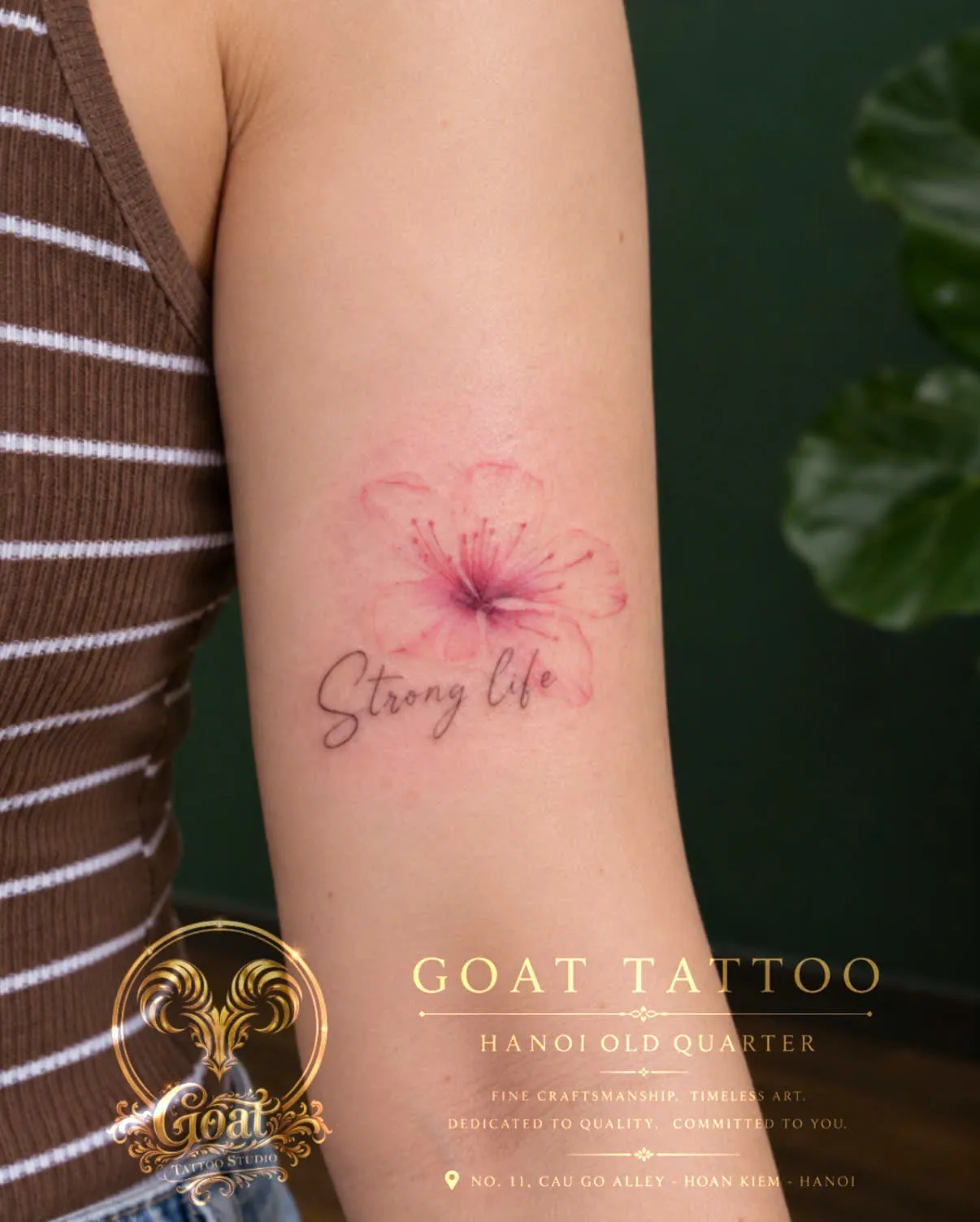 A pink cherry blossom tattoo with a Strong Life script underneath, on the upper arm, by GOAT Tattoo Studio in Hanoi's Old Quarter.