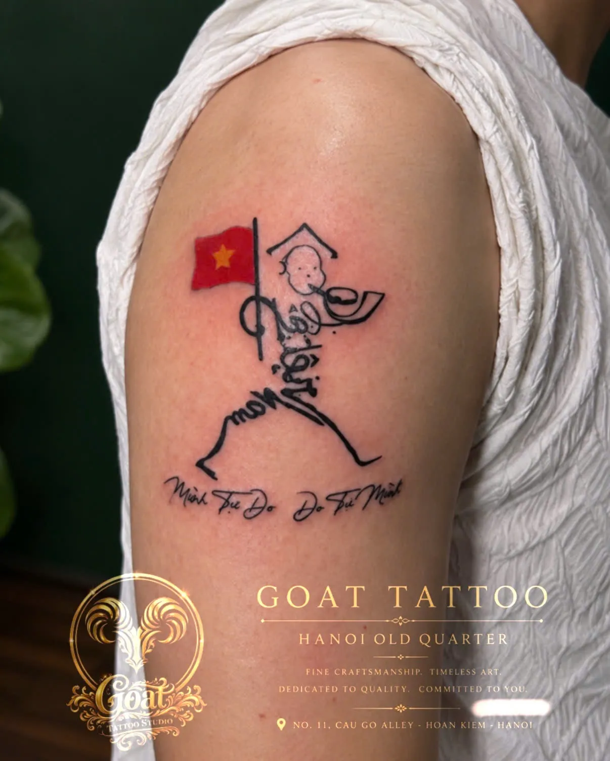 A calligram tattoo shaping a figure in a conical hat around Vietnamese script and a small flag, on the upper arm, by GOAT Tattoo Studio in Hanoi's Old Quarter.