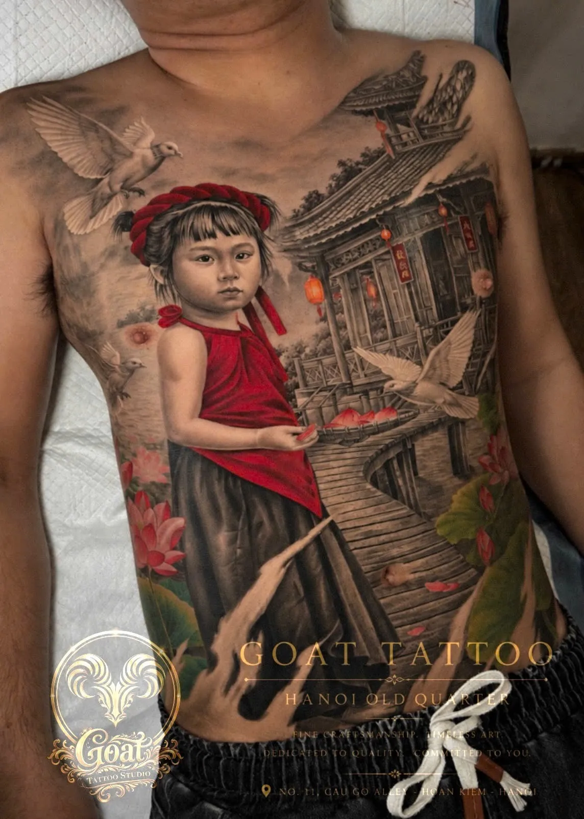 A full-color torso tattoo of a village girl on a lotus pond boardwalk, doves, and a lantern house, by GOAT Tattoo Studio in Hanoi's Old Quarter.